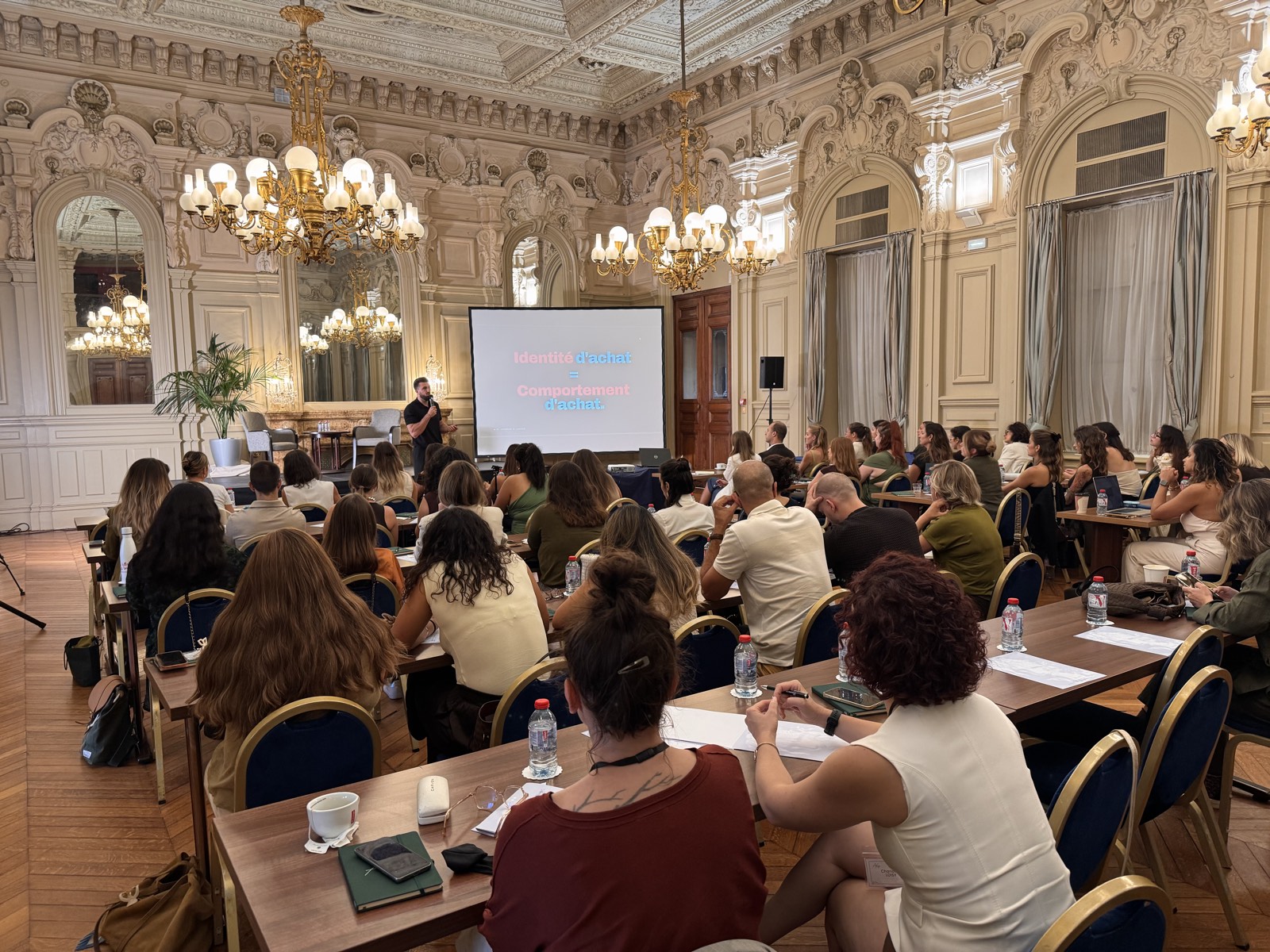 Une grande salle à moulures et lustres, pleine de participants assis face à l'écran pendant un événement de Julien.